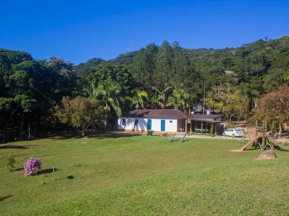 A casa branca de janelas azuis no fim do gramado, com a mata e a serra atrás