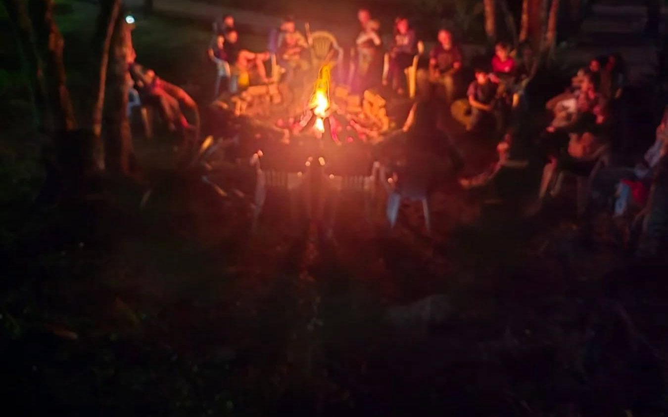 Roda de pessoas em volta da fogueira acesa, à noite, no meio das árvores