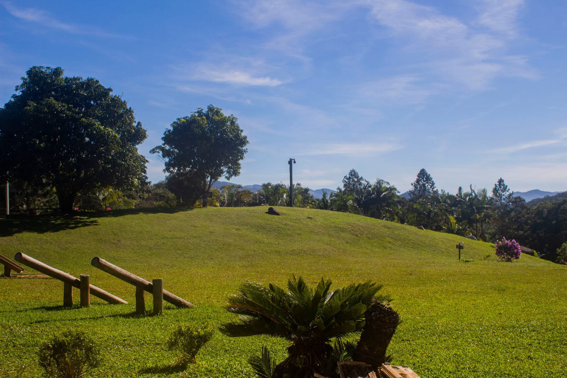 Gramado em morro com bancos de madeira, árvores e a serra ao fundo, debaixo de céu azul